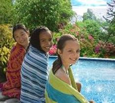 3 girls ssitting and wrapped in towels. In the background, a swimming pool surrounded by plants and trees.